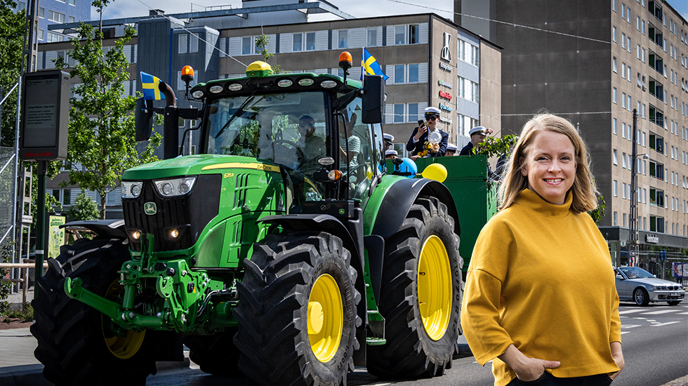 Leende kvinna framför grön traktor med svenska flaggor i stadsmiljö