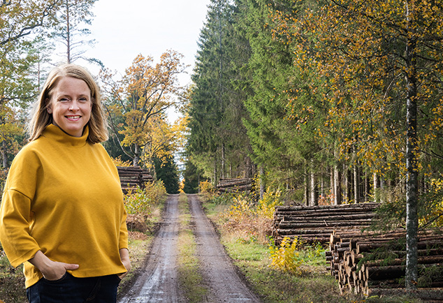 Leende kvinna i gul tröja står vid skogsväg med staplade timmerstockar