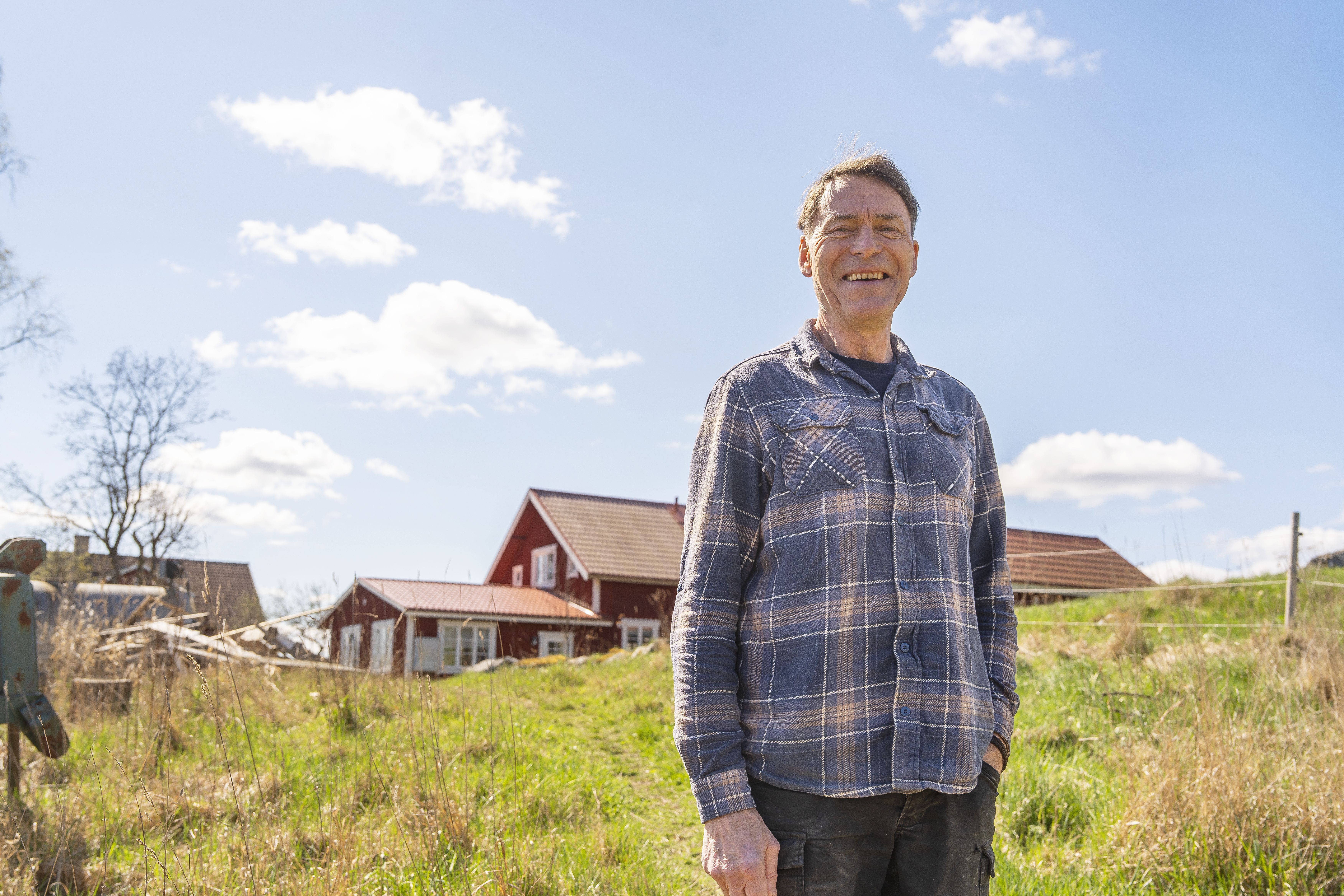 Leende man står på solig äng framför röda hus på landet