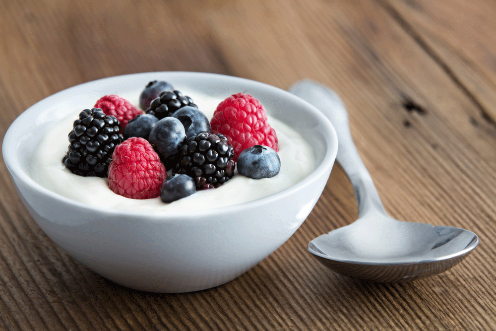 Skål med yoghurt toppad med färska hallon, blåbär och björnbär bredvid en sked