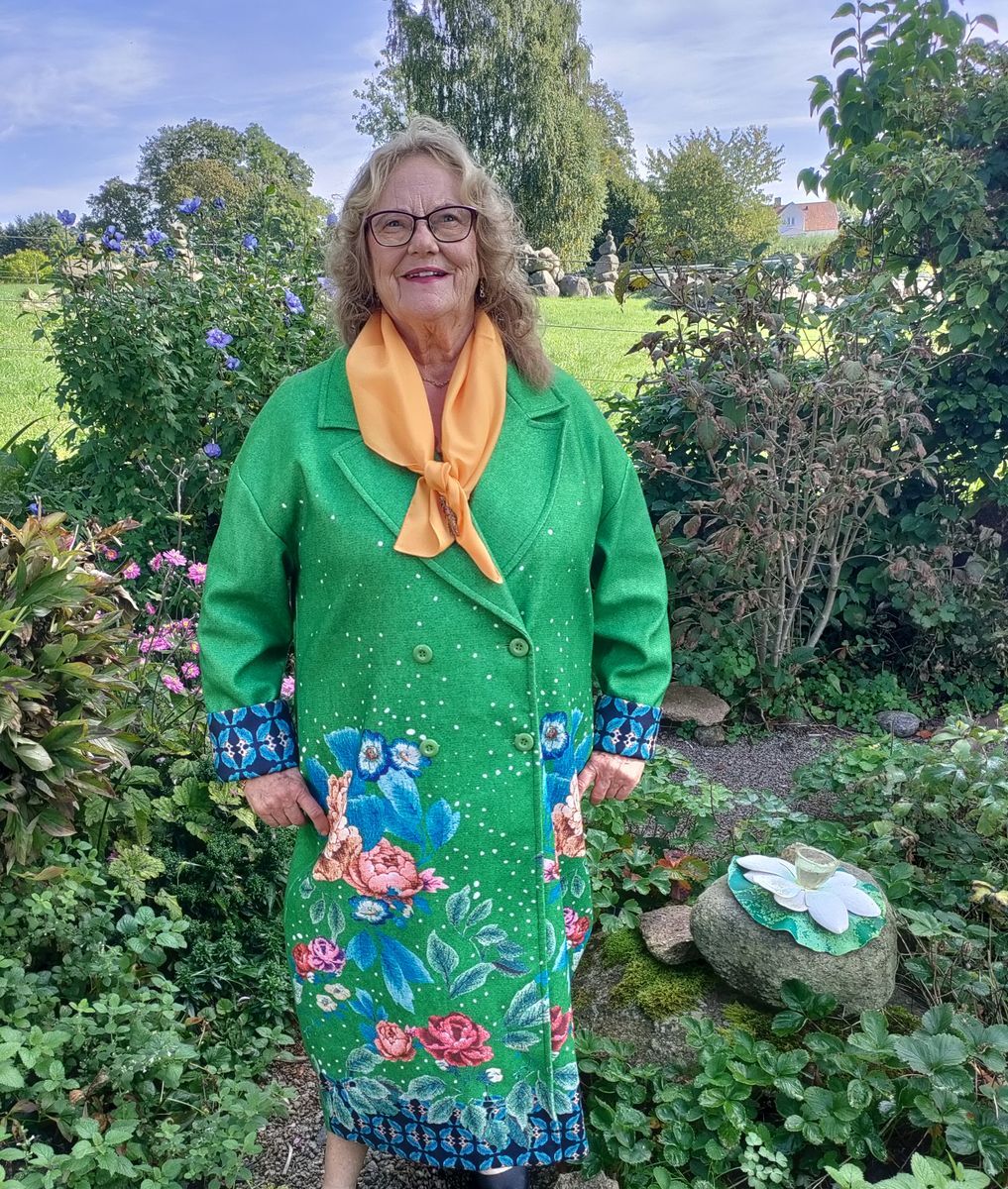 Ruth Hammarkvist i grön blommig kappa står leende i en prunkande trädgård.