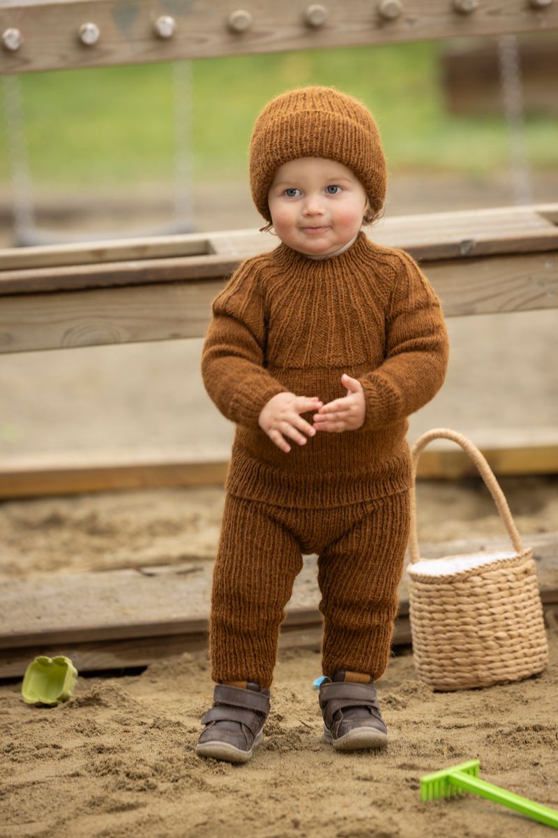 Litet barn i brun stickad outfit leker i sandlådan med korg bredvid sig
