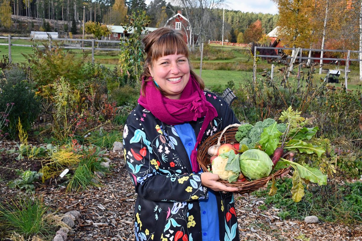 Leende Linda i trädgård håller en korg med nyskördade grönsaker på landet