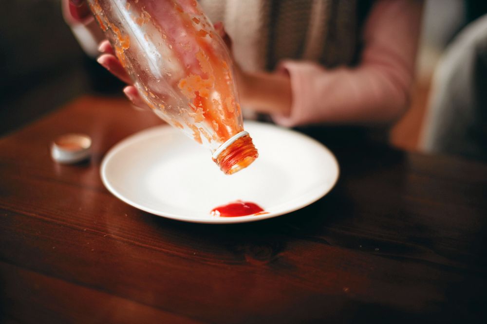 Person ur bild håller tom ketchupflaska upp och ner ovanför tallrik med en klick ketchup på.