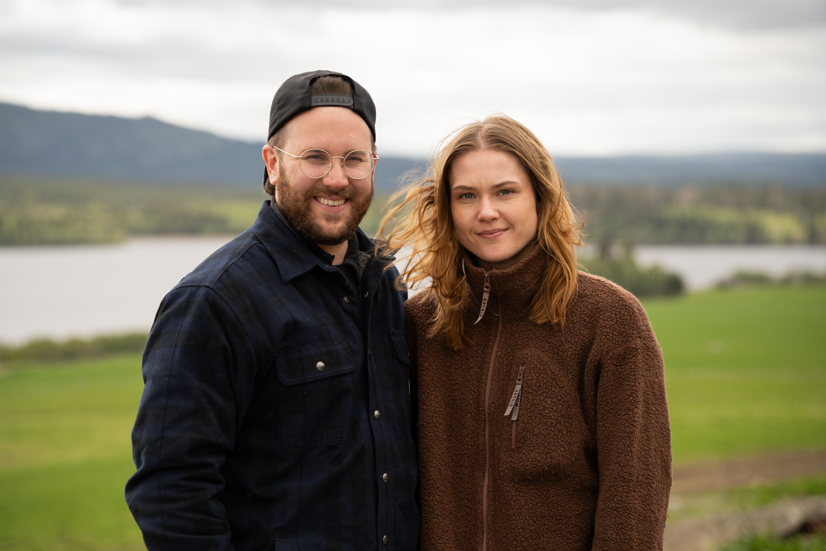 Två personer står nära varandra utomhus med sjö och berg i bakgrunden