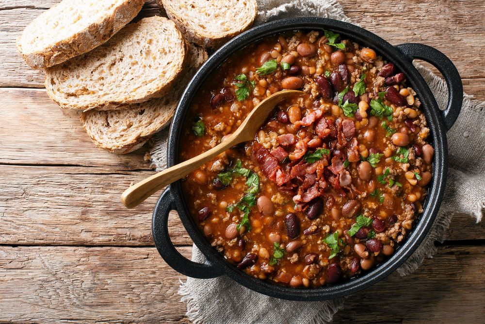 Cowboysoppa med bönor i svart gryta på handduk på träbord med uppskuret bröd intill.