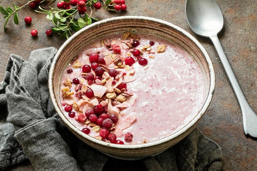Skål med rosa smoothiegröt toppad med lingon, nötter och frön på rustik bakgrund