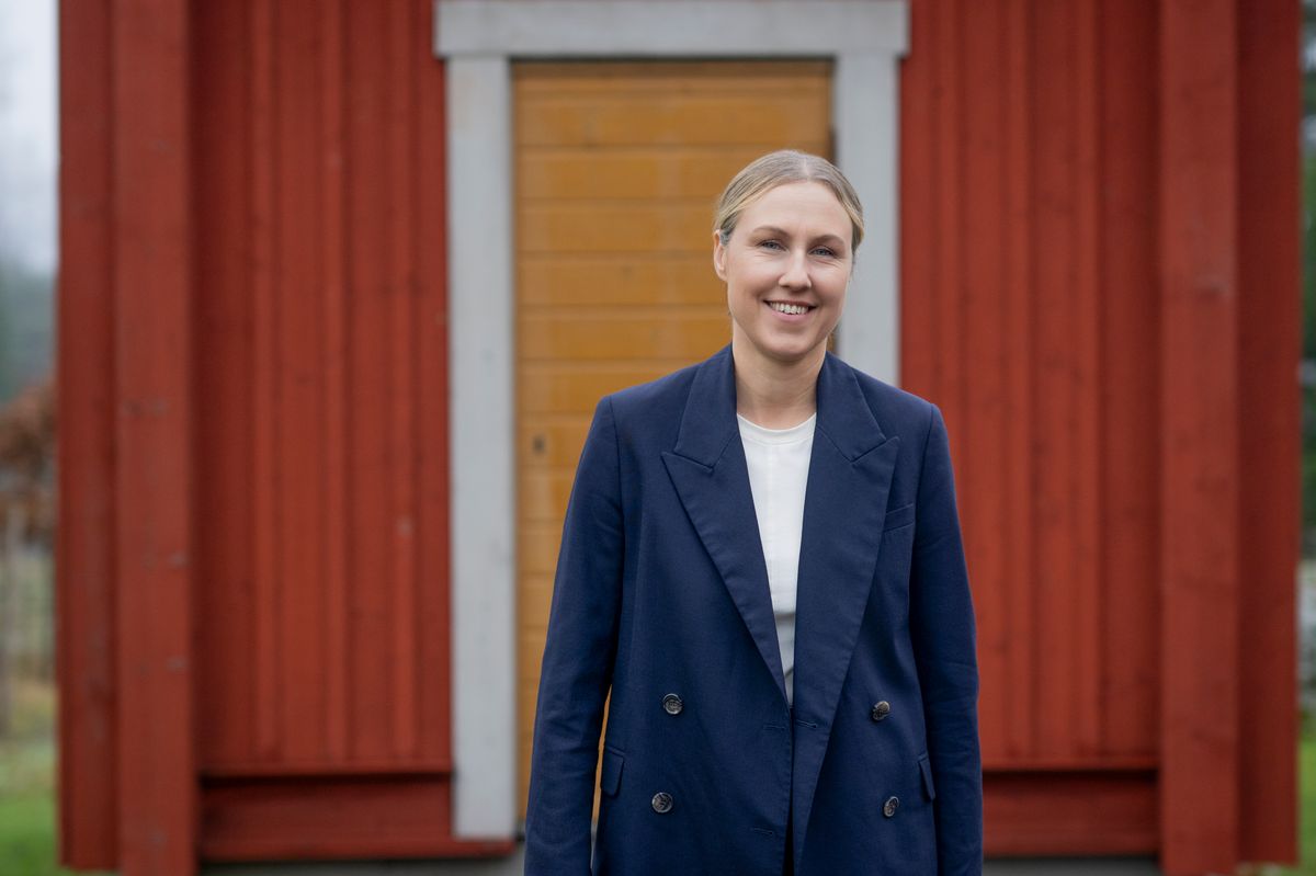 Leende person i mörk blå kavaj står framför en röd träbyggnad med gul dörr
