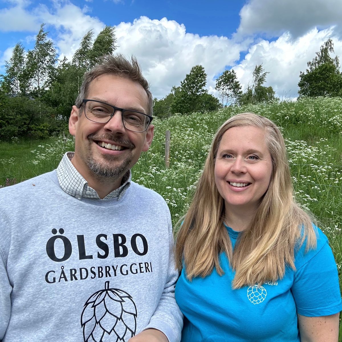 Rickard och Ylva Sjöberg står leende utomhus i grönskande landskap, med tröjor från gårdsbryggeri
