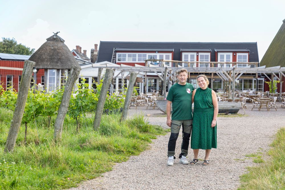 Arilds vingård. Paret Jonas och Anette Ivarsson står på grusgång framför lantlig restaurang med uteservering och vinrankor