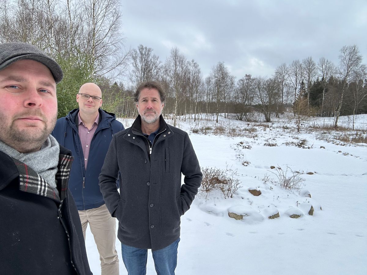 Tre män står utomhus på en snötäckt åker med träd i bakgrunden