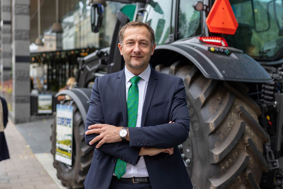Man i kostym med grön slips står framför en stor traktor i stadsmiljö
