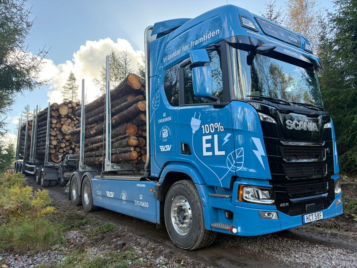 Blå eldriven lastbil transporterar timmer på grusväg i skogsmiljö