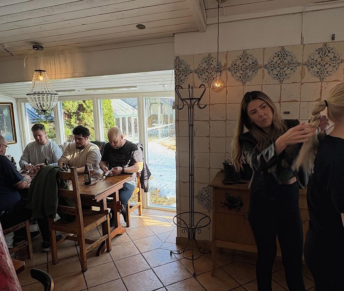 Grupp vid träbord nära fönster, kvinna i förgrunden fixar hår på annan person