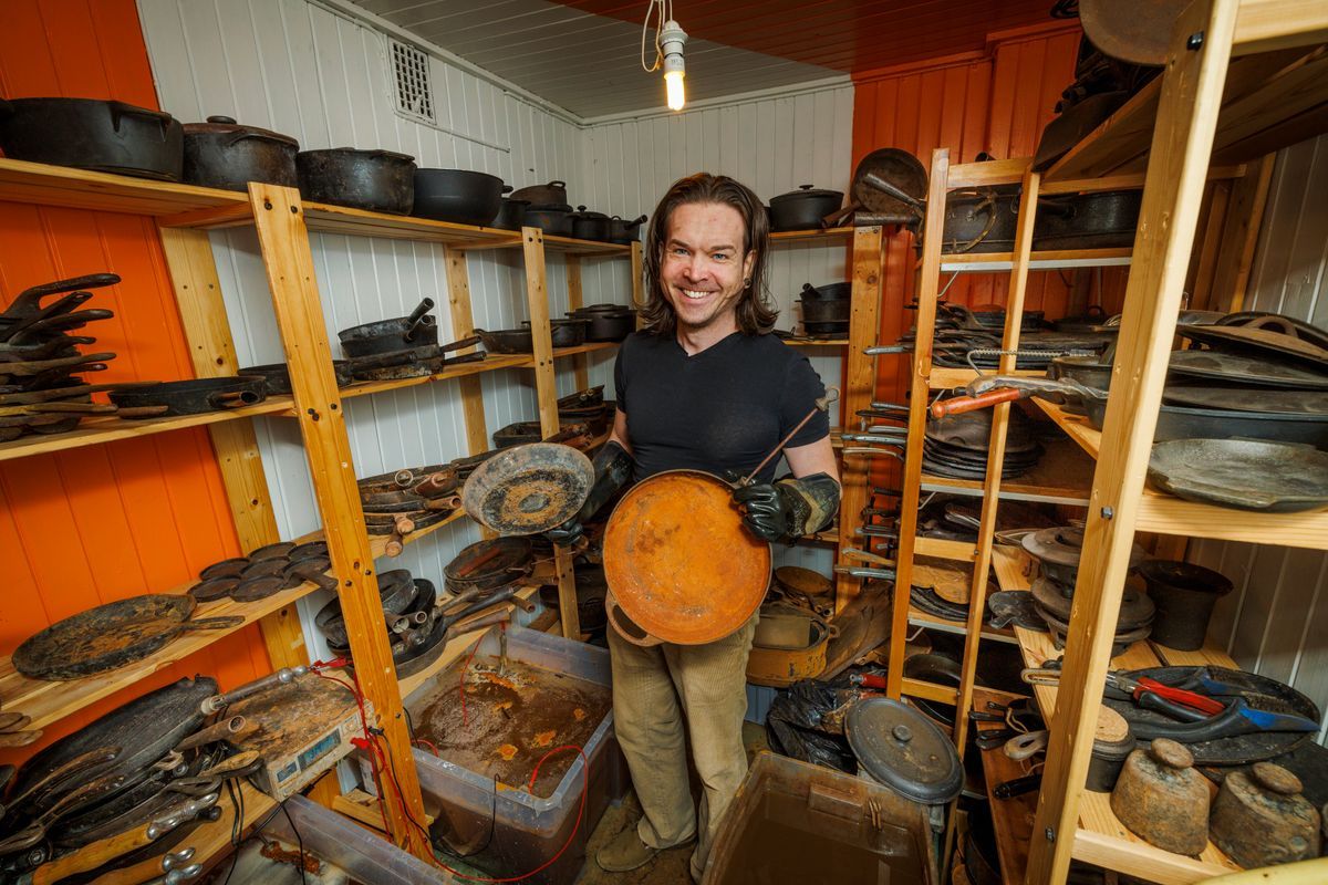 Leende Joel Bergqvist i förråd fullt av gjutjärnspannor och grytor, håller en rostig panna