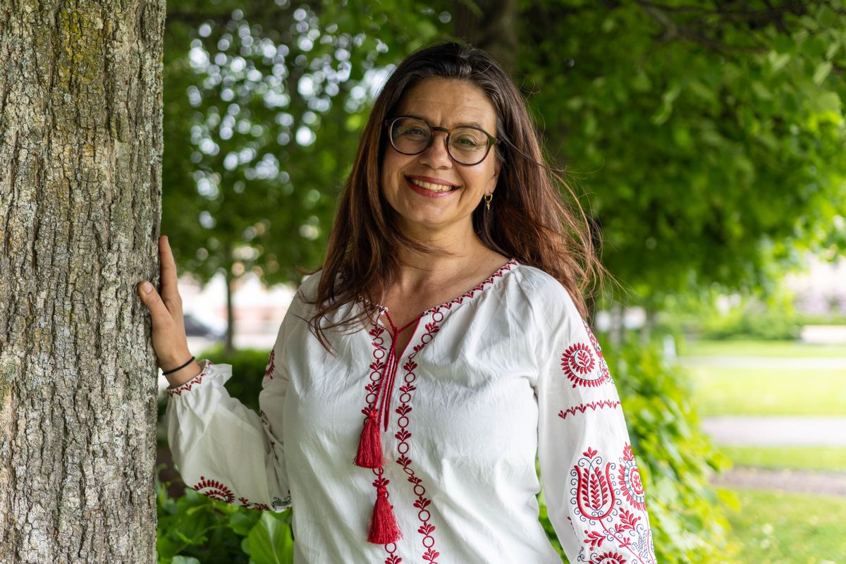 Leende kvinna med glasögon i vit broderad blus står vid ett träd i parkmiljö