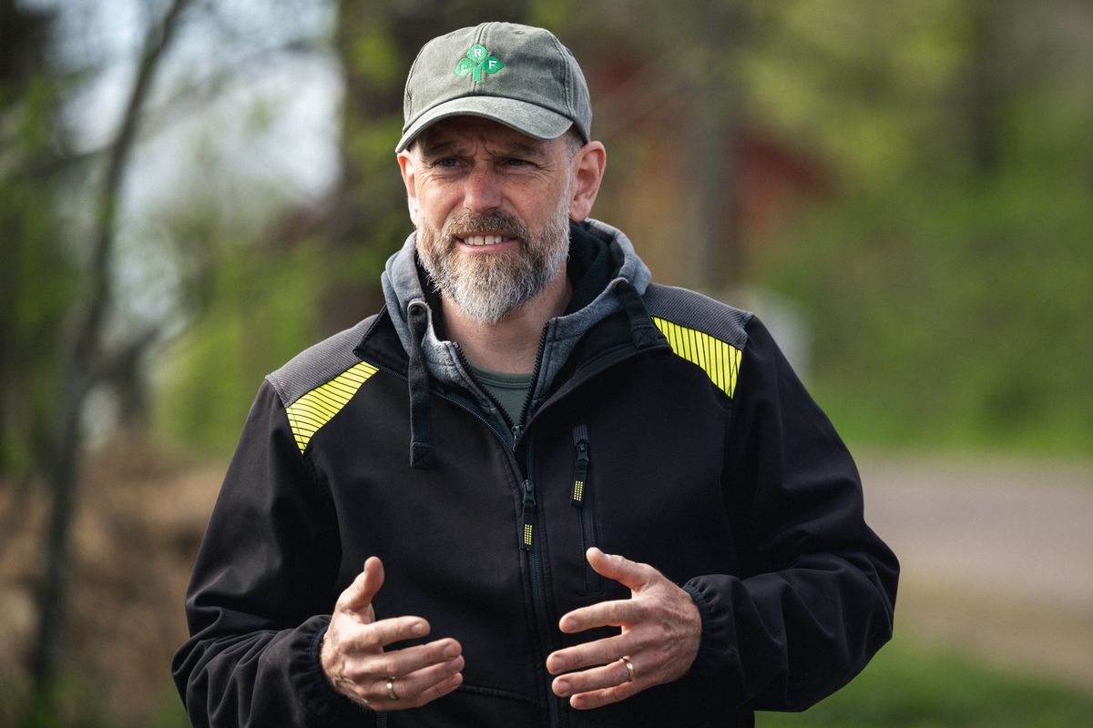 Skäggig man med keps och jacka gestikulerar utomhus i naturmiljö