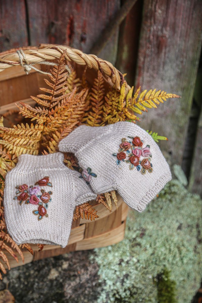 Stickade plagg med broderade blommor i korg med höstbräken mot trävägg