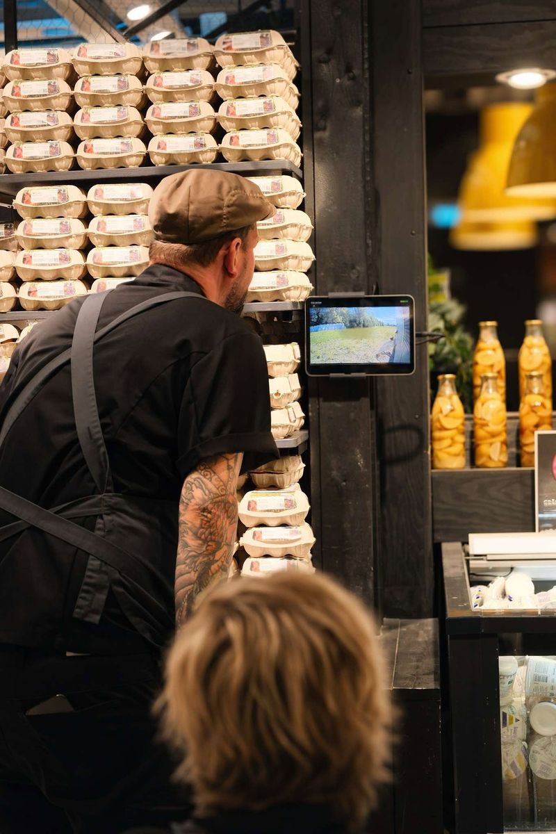 Butiksanställd tittar på surfplatta där hönsen livestreamas, bredvid staplade äggkartonger i butik