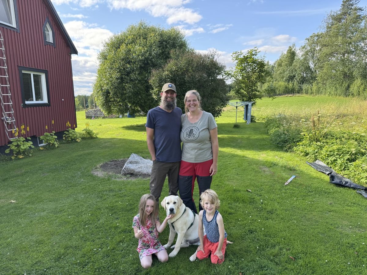 Familj med två barn och hund poserar på gräsmatta vid rött hus på landet