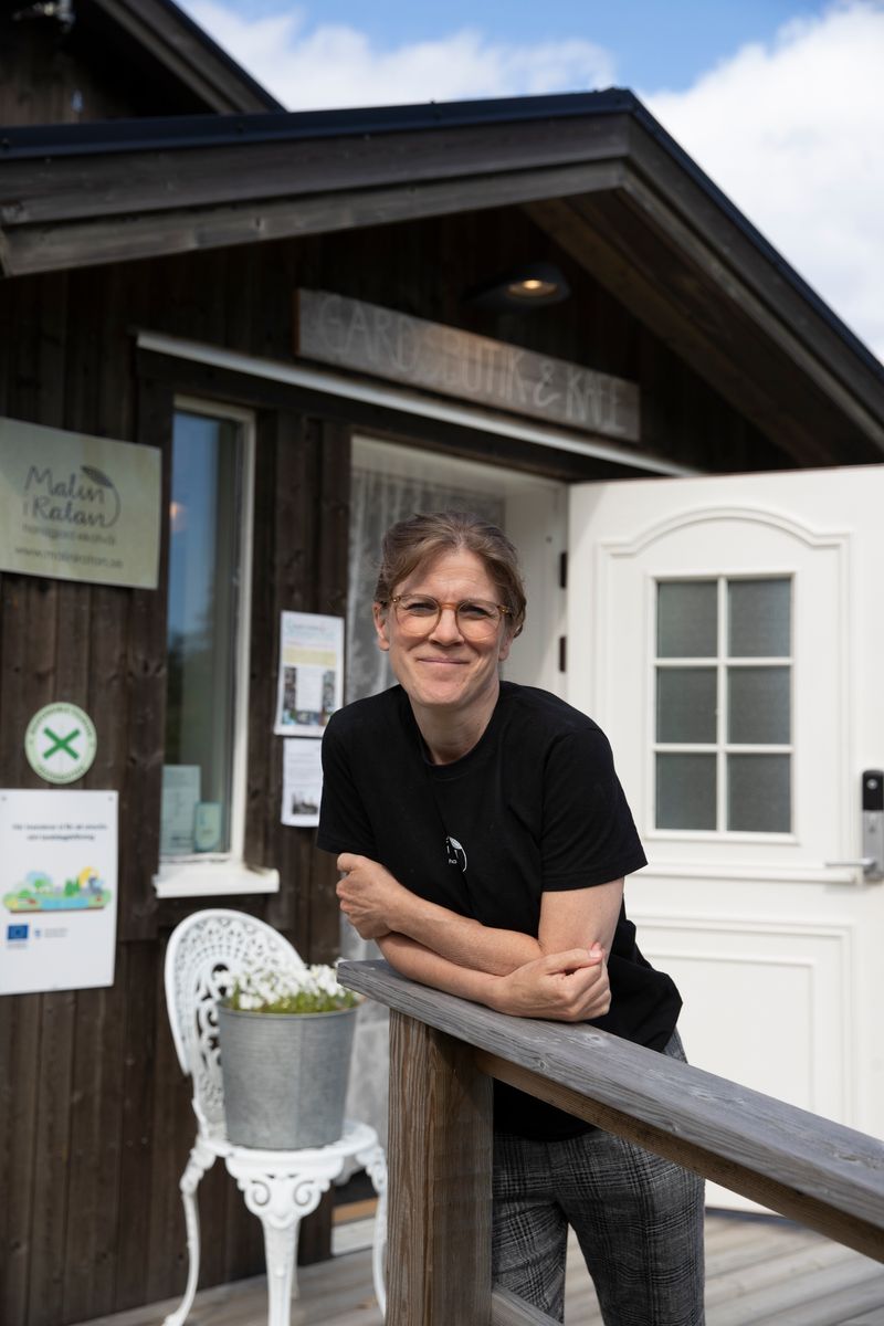 Leende Malin Lindmark lutar mot räcke utanför en träbyggnad med vit dörr och skylten Malin i Ratan