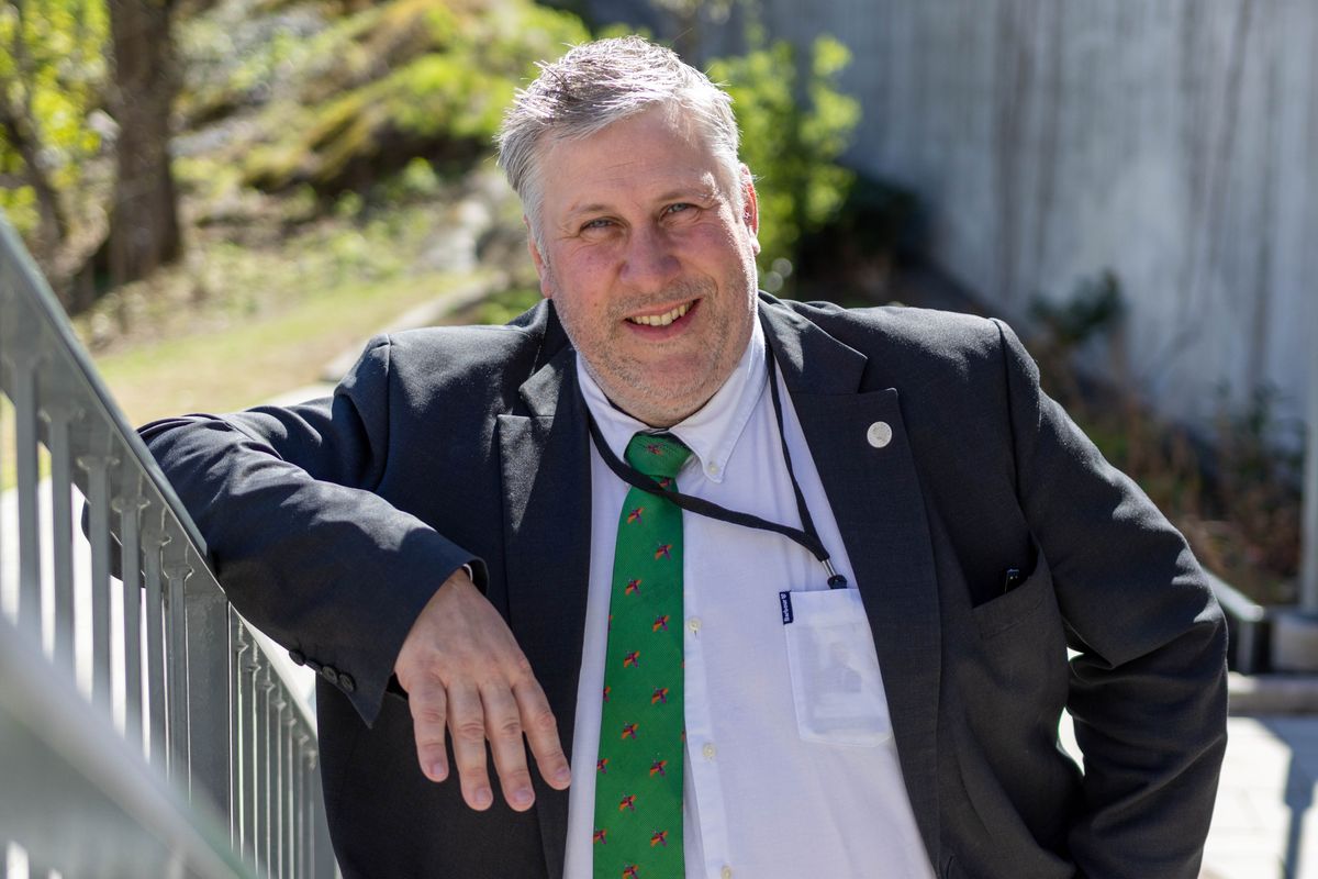 Leende man i kostym och grön slips lutar sig mot ett räcke utomhus en solig dag