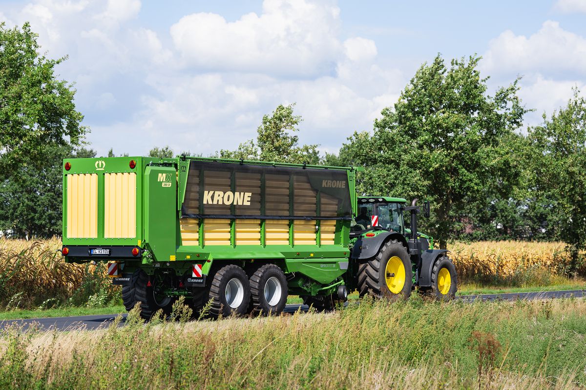 Grön traktor med Krone-vagn kör på landsväg bredvid åker och träd