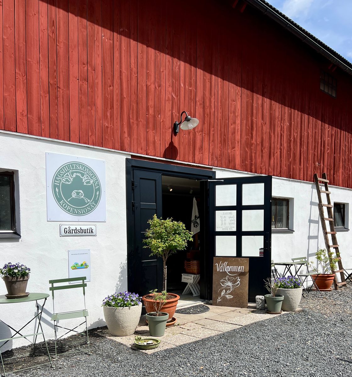Ingång till gårdsbutik i röd ladugård, med krukväxter och utemöbler framför.
