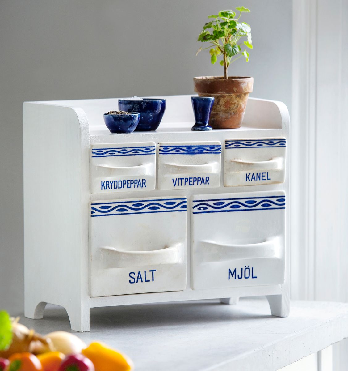 Vit kryddbyrå med blå detaljer, märkta lådor för salt, mjöl och kryddor, liten krukväxt ovanpå