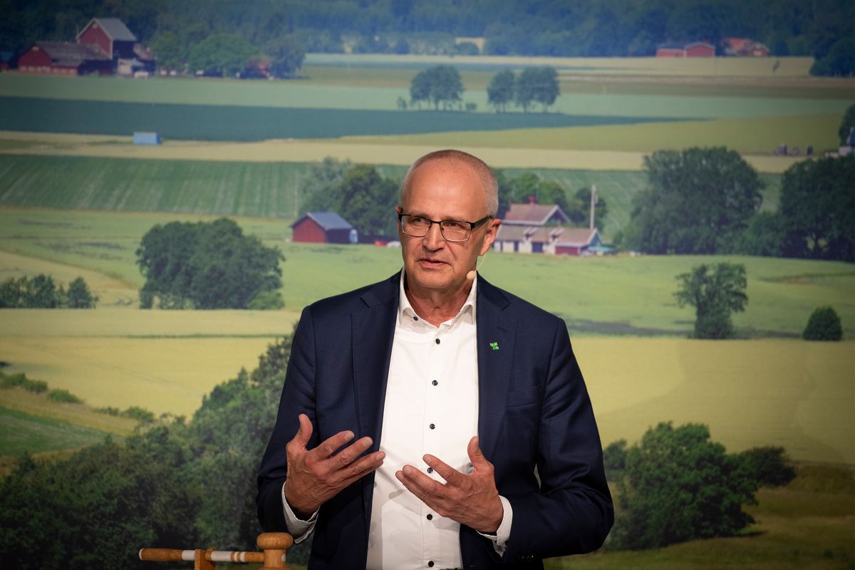 Man i kostym talar framför en lantlig bakgrund med åkrar och gårdar.
