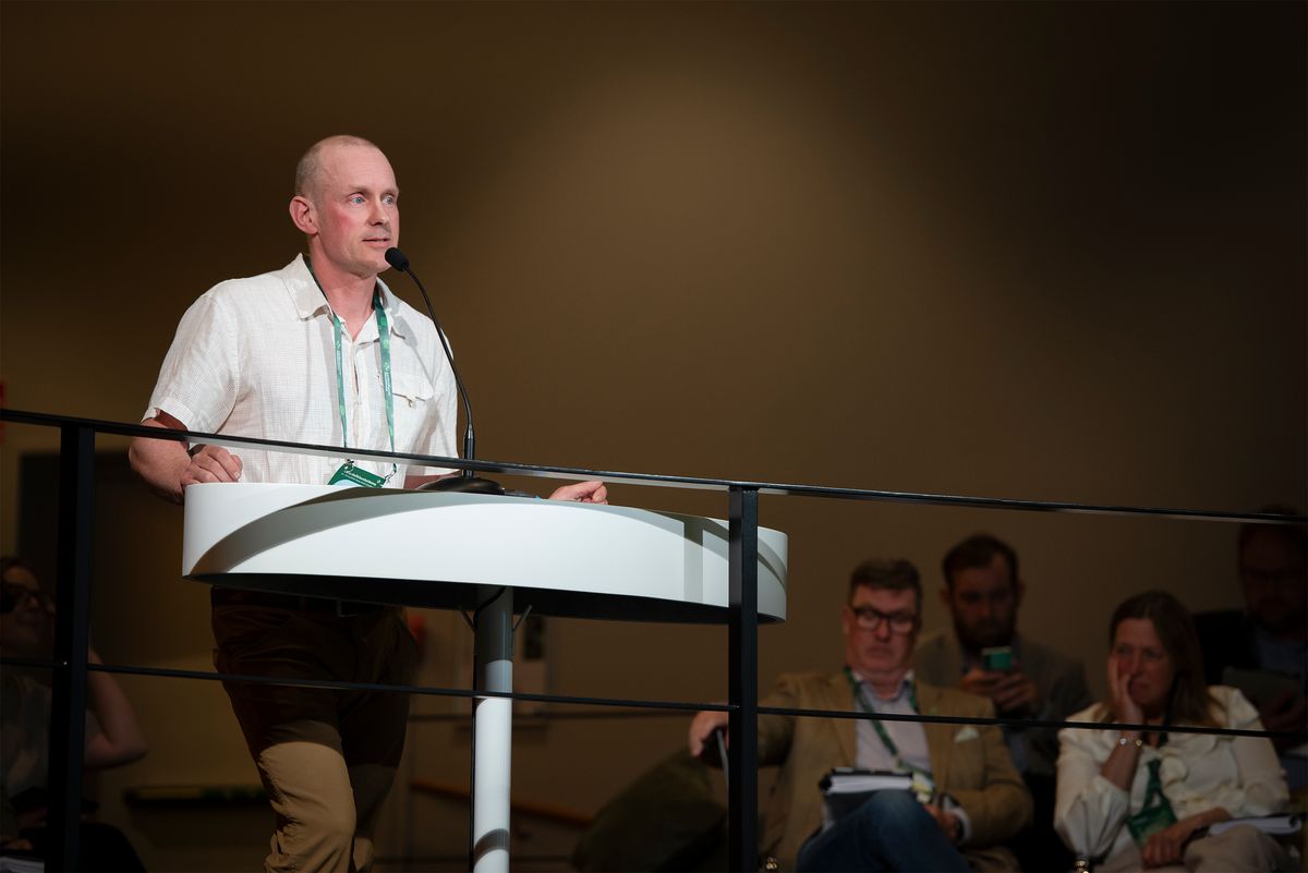 Man talar vid podium på konferens medan åhörare lyssnar i bakgrunden