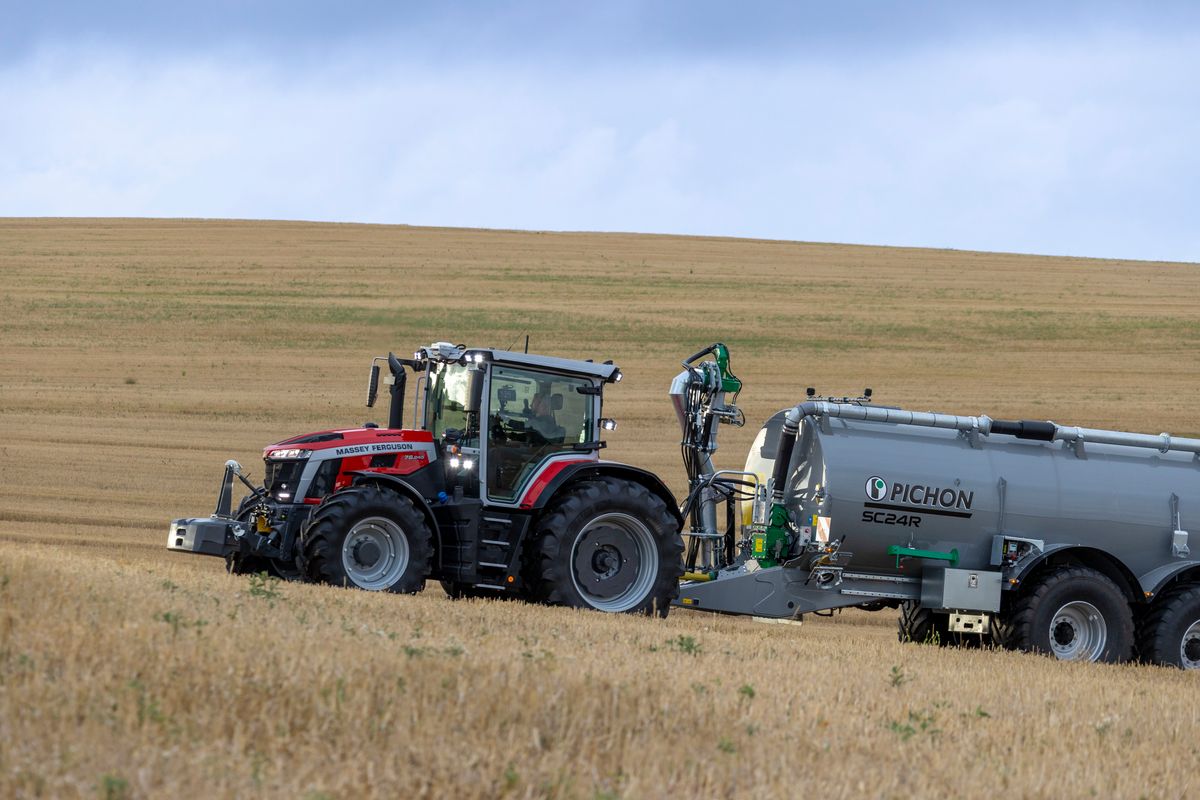 Traktor med stor gödseltank kör över en gulbrun åker under mulen himmel