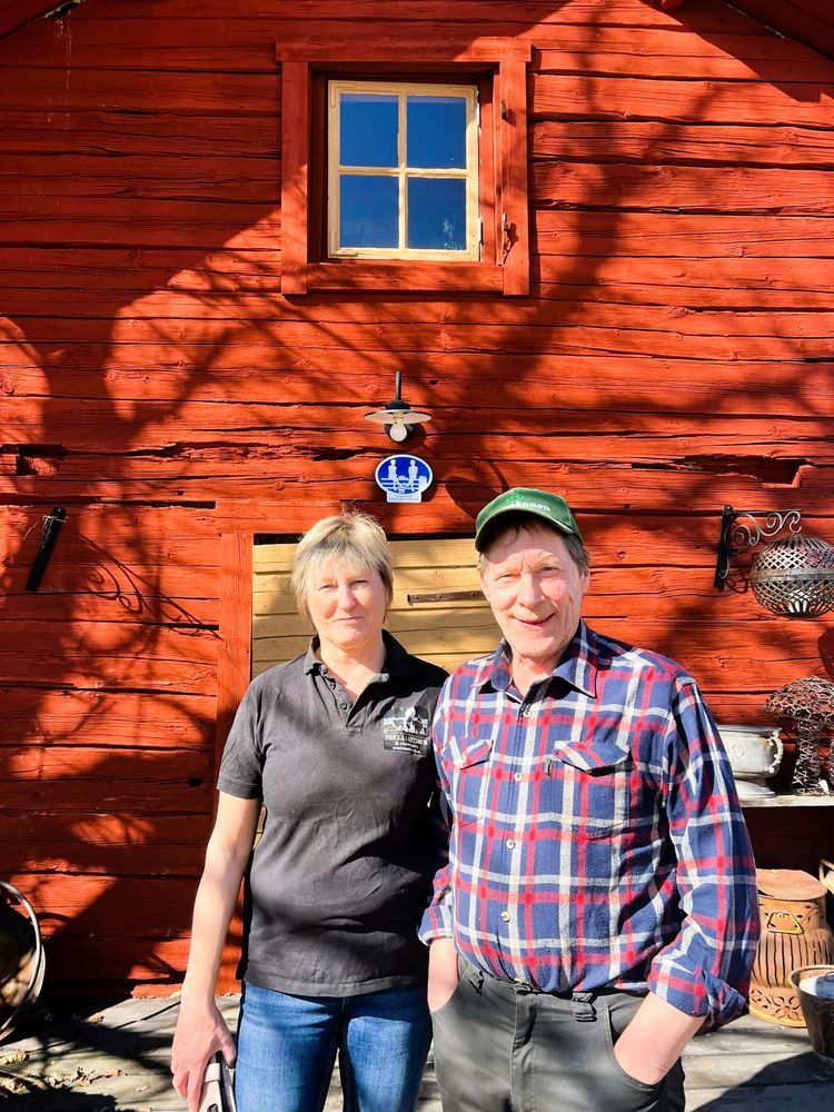 Åsa och Per Pesula personer framför en röd träbyggnad i solsken.