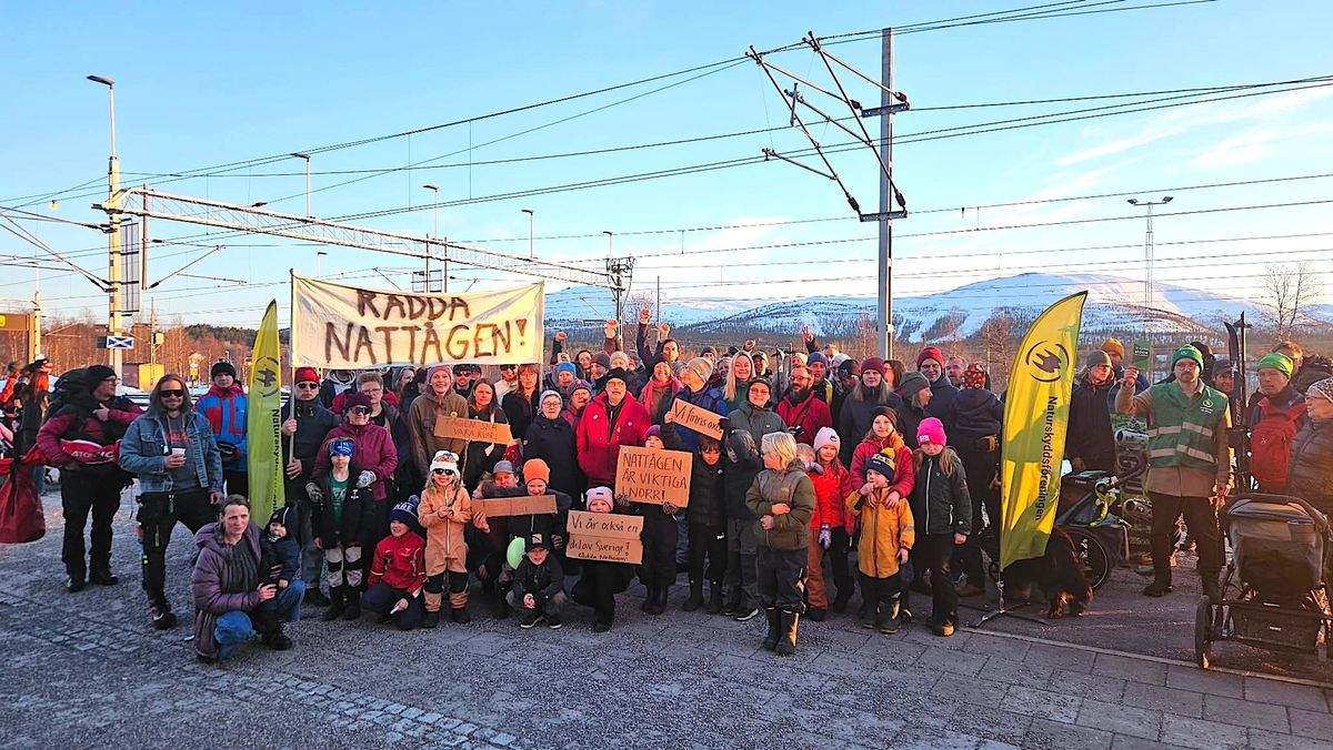 Stor grupp människor demonstrerar vid järnväg med banderoll ”Rädda nattågen!” och berg i bakgrunden.
