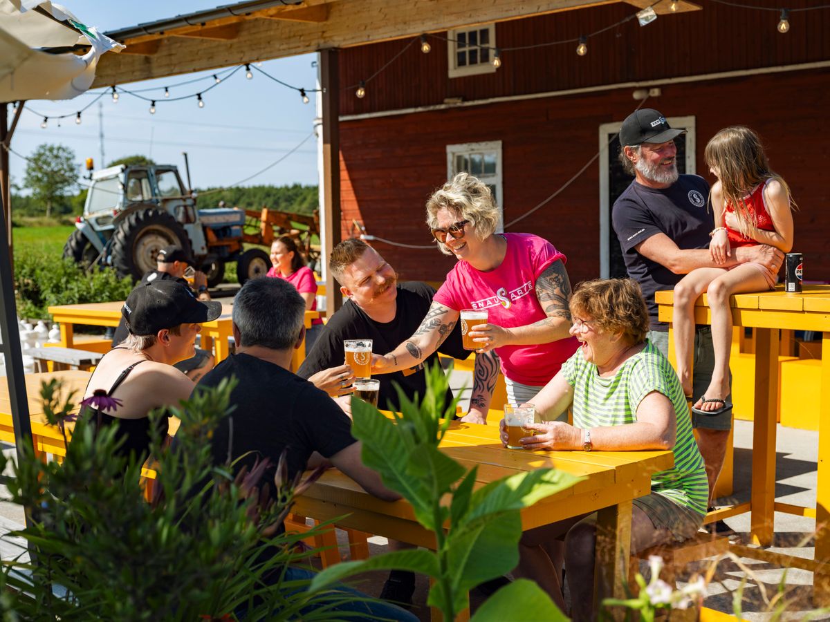 Vuxna och barn fikar vid gula bord på gårdscafé en solig sommardag