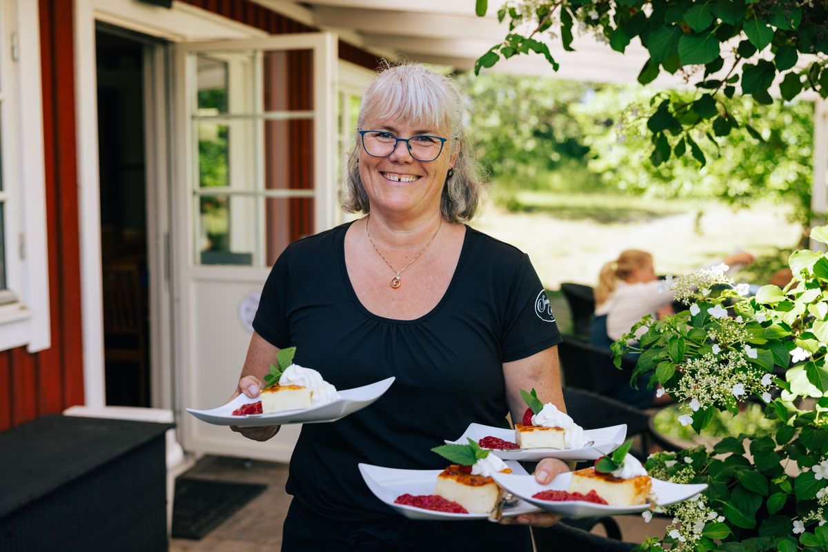 Leende kvinna utanför röd trävilla, håller flera tallrikar med bitar av ostkaka på, på Sussies café.