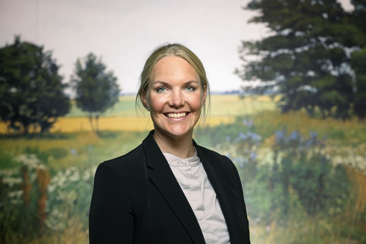 Leende kvinna i kavaj framför en somrig landskapsbakgrund med träd och åkrar
