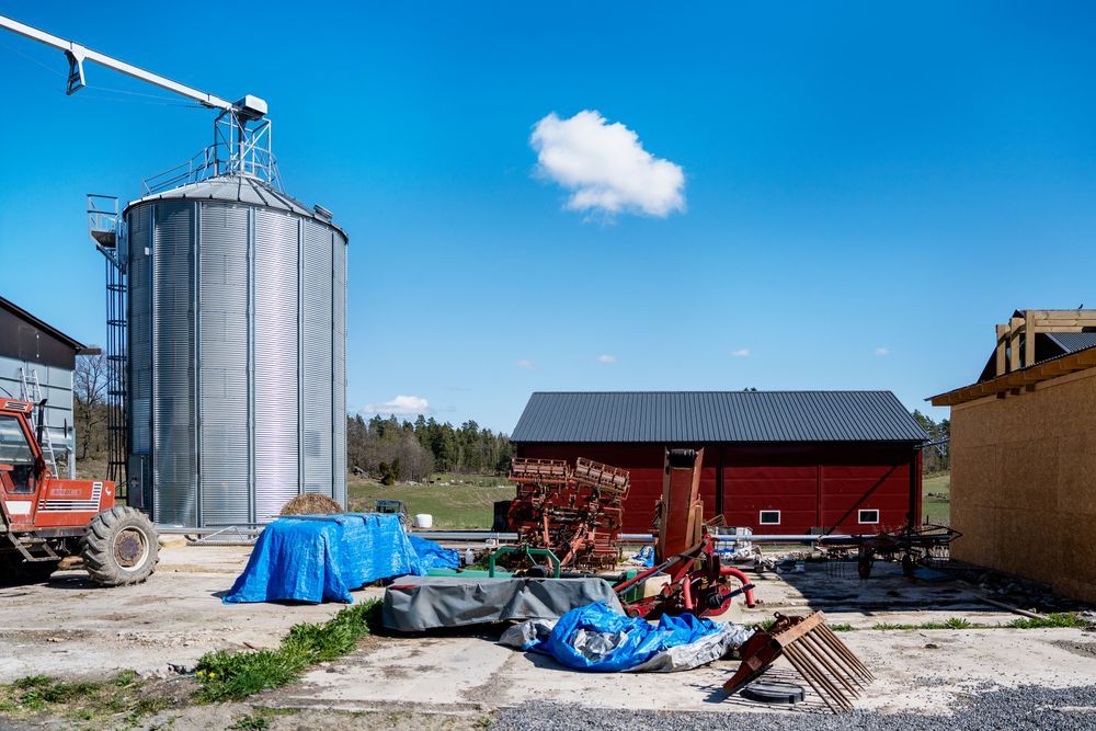 Gård med metallsilo, röda lador och utspridda jordbruksmaskiner under blå himmel