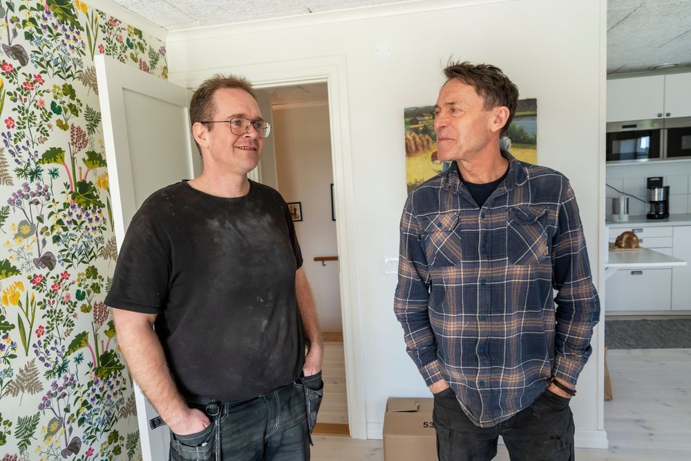 Osku Varis i svart t-shirt och Thomas Björklund i arbetskläder samtalar i ett ljust hem med blommig tapet