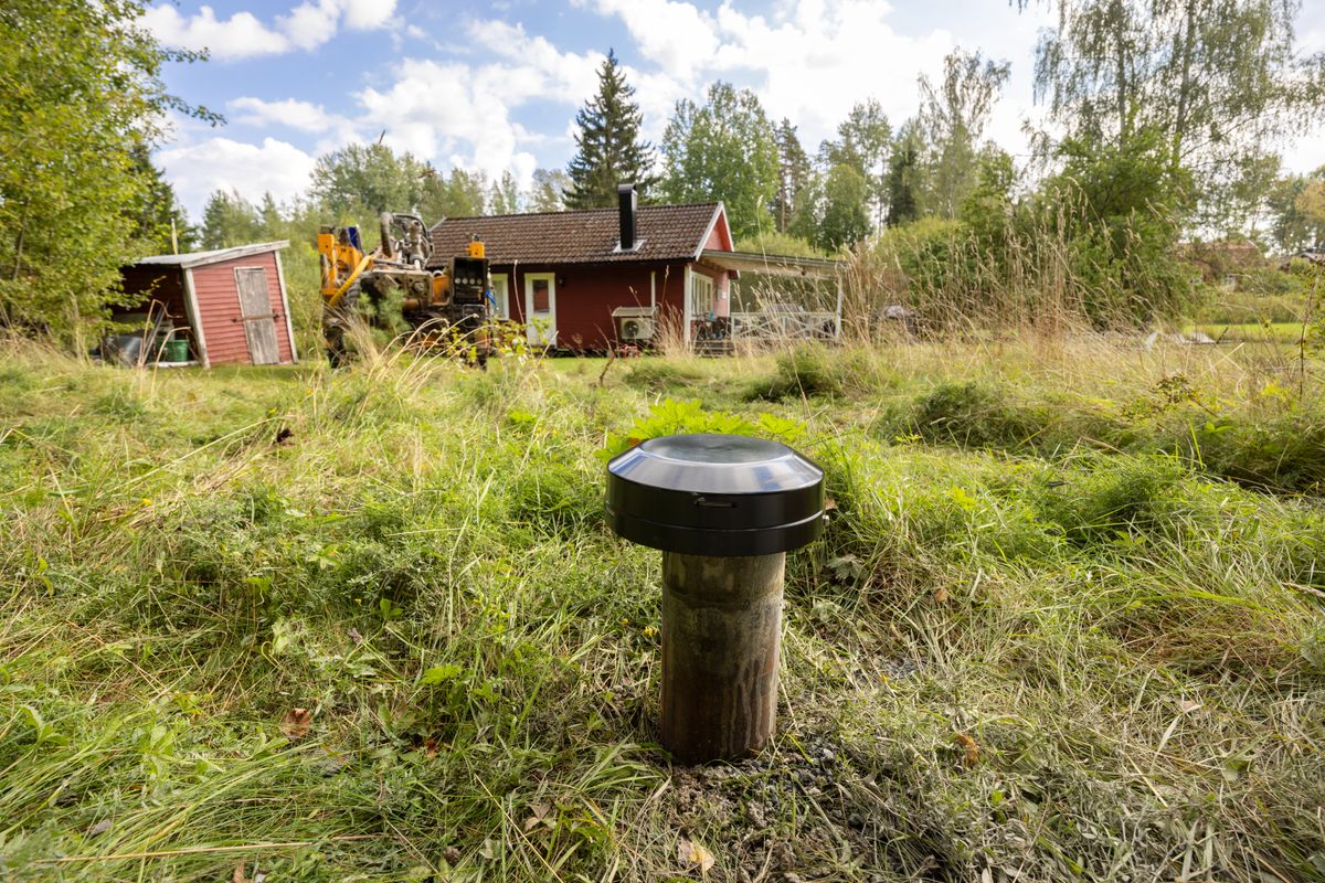 Brunnslock i gräset framför röd stuga med grävmaskin i bakgrunden