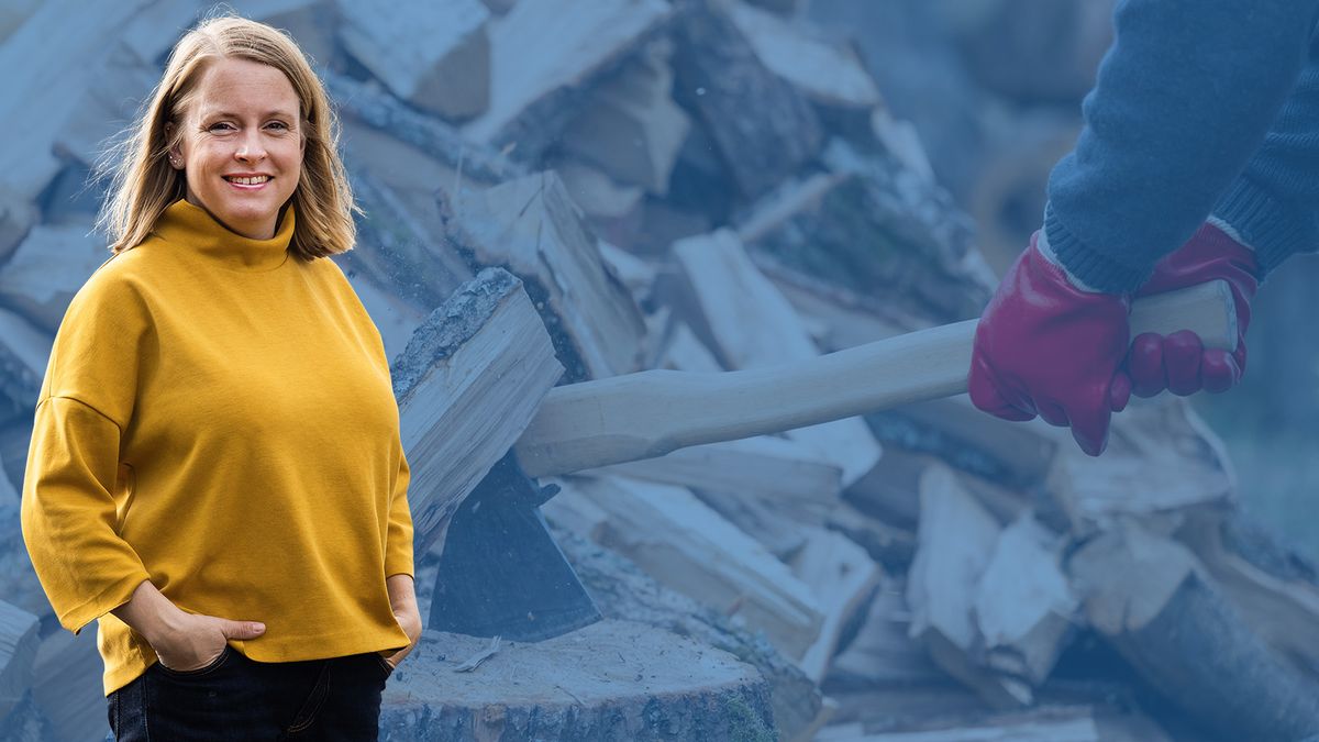Leende Anna Nilsson i gul tröja framför huggen ved och en yxa som slår i en träklabbe