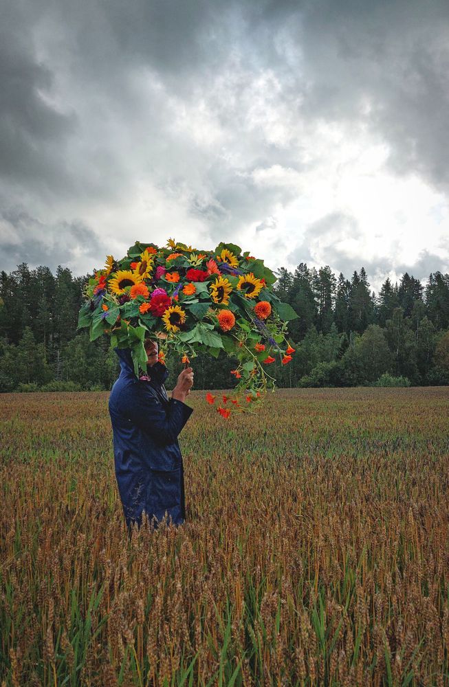 Kvinna på lite avstånd i en åker som står och håller paraply gjort av blommor.
