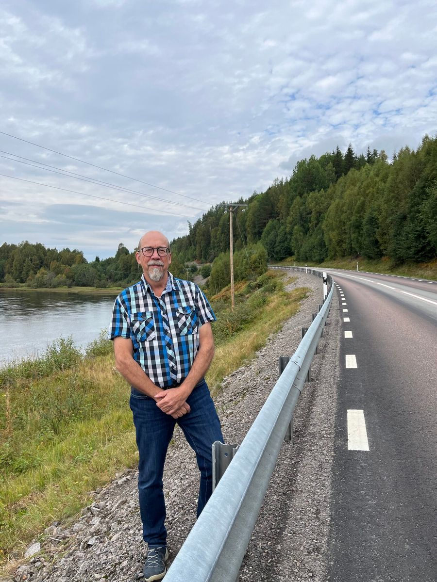 Jan-Anders Carlsson i rutiga kläder står vid landsväg intill en lugn flod och skog