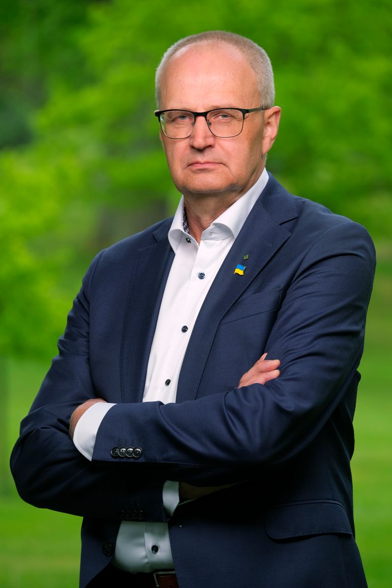 Äldre man i kostym med korslagda armar utomhus framför grön bakgrund