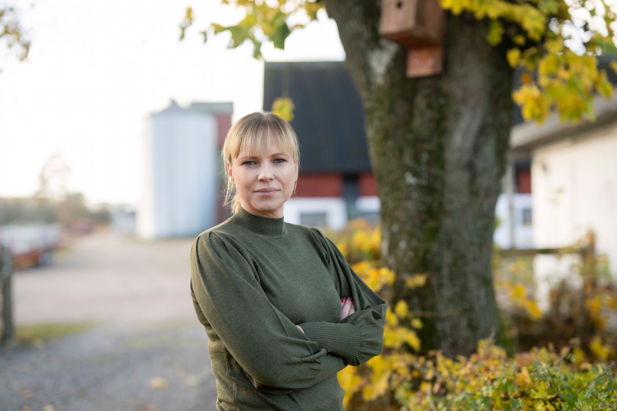 Kvinna i grön tröja står vid ett träd på en lantlig gård med höstlöv