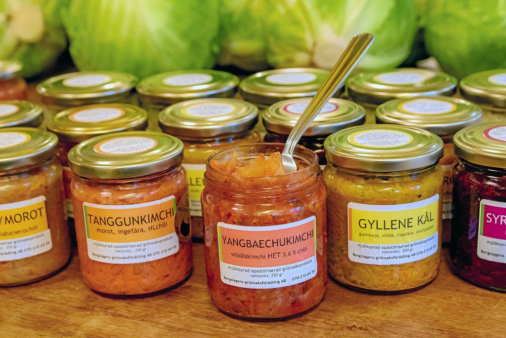 Glasburkar med olika sorters kimchi och syrad kål på en träbänk framför kålhuvuden