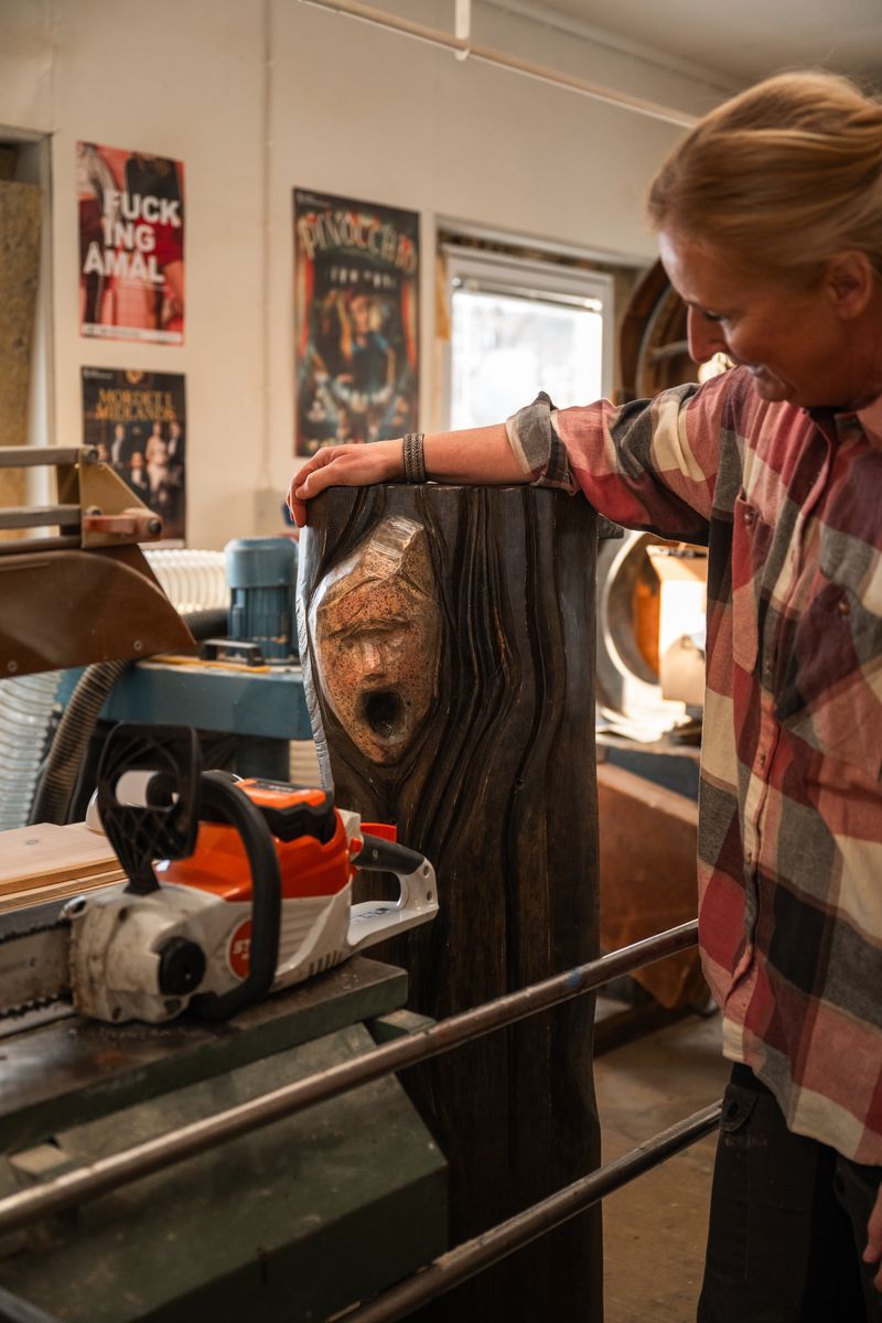 Eva-Karin i verkstad med motorsåg, arbetar på träskulptur med ansikte och öppet gap
