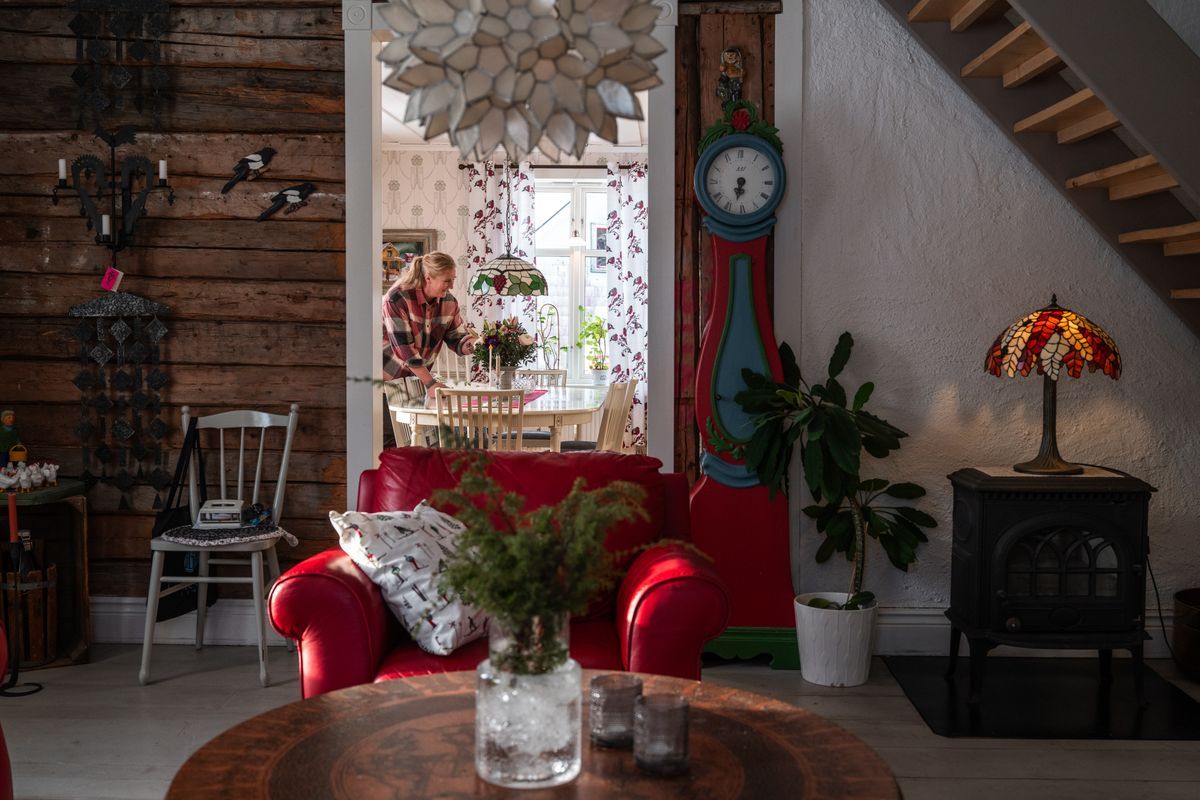 Mysig lantlig vardagsrumsmiljö med röd fåtölj och Eva-Karin vid matbord i bakgrunden