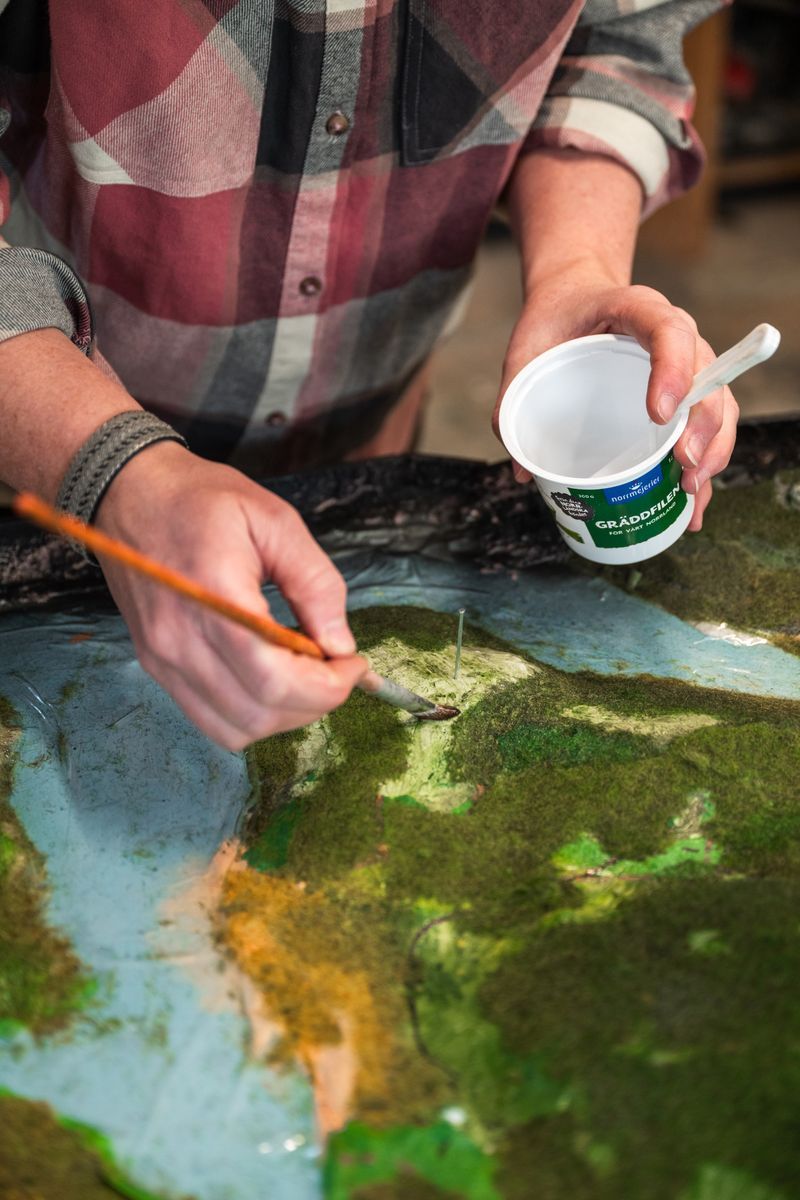 Eva-Karin målar en landskapsmodell med pensel och håller en yoghurtbägare i andra handen