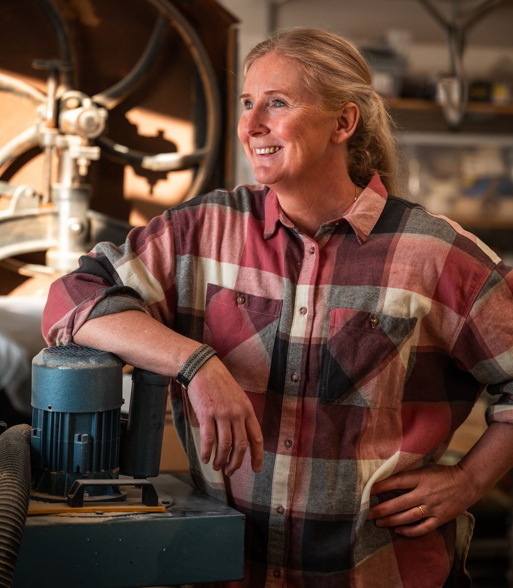 Leende Eva-Karin Fröberg i flanellskjorta lutar sig mot en maskin i snickeriverkstad